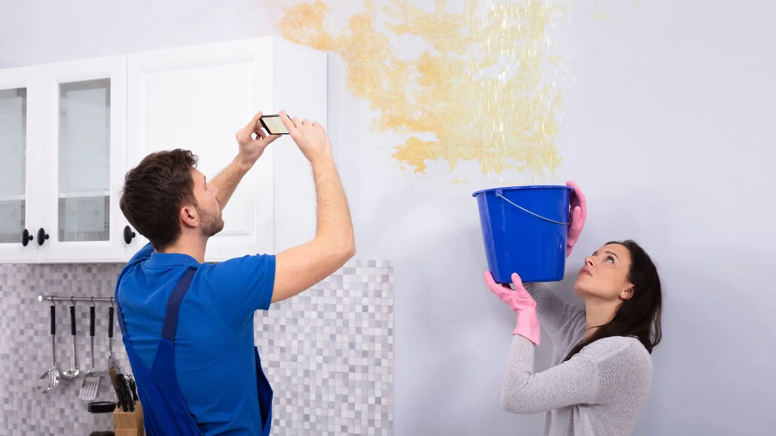 Homeowner photographing a ceiling stain while another person catches drips in a bucket